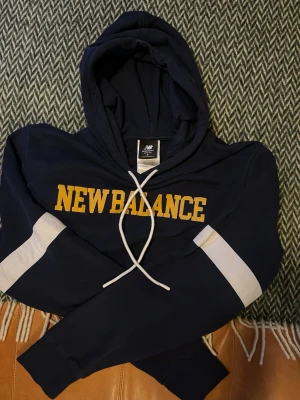 Mörkblå New Balance hoodie - Snygg mörkblå hoodie från New Balance med gul logga på bröstet och vita snören i huvan. Ärmarna har vita detaljer som ger en sportig vibe. Perfekt för dig som gillar streetwear och vill ha något bekvämt och stilrent.