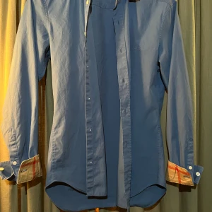 Blå skjorta från Burberry med rutiga detaljer - Snygg blå skjorta från Burberry med klassiska rutiga detaljer på insidan av manschetterna. Skjortan har lång ärm, knappar framtill och är tillverkad i bomull. Perfekt för dig som gillar stilrena plagg med en twist.