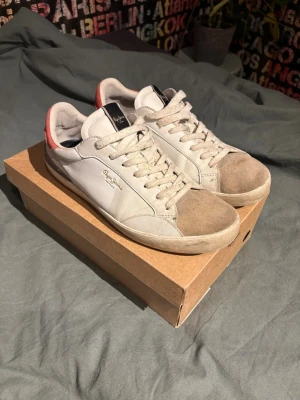Vita sneakers från Pepe Jeans - Snygga vita sneakers från Pepe Jeans med beige mockadetaljer på tån och röd detalj vid hälen. Skorna har klassisk snörning och rund tå. Perfekta för en avslappnad och stilren look. Storlek 43. Billigare vid snabb affär. Tvättas innan de fraktas