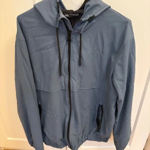 Blå vindjacka med huva från Zara - Snygg blå vindjacka från Zara med huva och svarta dragsnören. Jackan har hel dragkedja framtill, två fickor med dragkedja och elastiska muddar vid ärmar och nederkant. Perfekt för blåsiga dagar och enkel att matcha med streetwear.