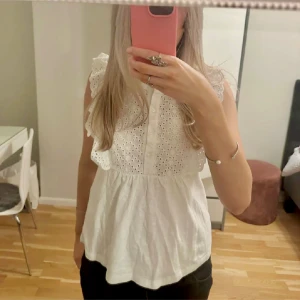 Vit ärmlös blus från JDY XS - Supersöt vit blus från JDY i storlek XS. Blusen är ärmlös med volangdetaljer på axlarna och har broderade hålmönster framtill. Knäppning med små knappar framtill och lätt utställd nertill. Perfekt för en fräsch och somrig look! Lite sminkfläckar på kragen se bild 5!