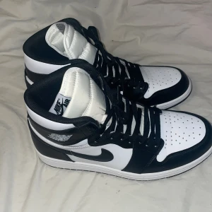 Nike Air Jordan 1 svart/vit sneakers - Säljer ett par Nike Air Jordan 1 sneakers i svart och vitt. Klassisk high-top modell med svart swoosh, perforerad tåbox och snörning. Skorna har en stilren kontrast mellan svart och vitt skinn, samt ikoniska Air Jordan-loggan på sidan. Perfekta för dig som gillar streetwear.