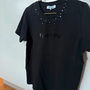 Svart t-shirt med stenar från Miu Miu - Svart t-shirt från Miu Miu med glansiga svarta stenar runt halsringningen och diskret logga framtill. Klassisk passform och korta ärmar. Tillverkad i mjuk bomull för en skön känsla. Perfekt för dig som vill ha en lyxig och stilren look. Storlek xl men uppskattar den till en m/l. Jag har användt den två ggr och märket sitter därför inte längre på men jag har kvar det. Köpt i Danmark för 900 DKK äkta 