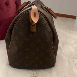 Louis Vuitton speedy 35  - Säljer en klassisk Louis Vuitton resväska i brunt med ikoniskt monogrammönster och detaljer i ljust skinn. Väskan har robusta handtag, dragkedja och lås med Louis Vuitton Paris-ingravering. Perfekt för dig som vill resa med stil.
