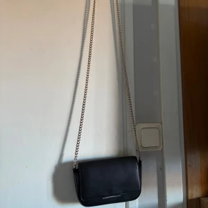 Svart axelväska från H&M med kedja - Stilren svart axelväska från H&M med silverfärgad kedjerem. Väskan har ett minimalistiskt uttryck med en liten metall-detalj på locket och magnetlås. Perfekt storlek för det viktigaste och enkel att matcha till olika outfits.