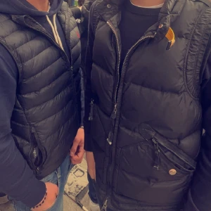 Svart dunväst från Parajumpers - Säljer en svart dunväst från Parajumpers med hög krage och dragkedja framtill. Västen har två snedställda fickor med dragkedja och knapp, samt en ikonisk gul detalj vid halsen. Perfekt att slänga över en hoodie eller tröja för en snygg lager-på-lager-look. Nypris 6049kr