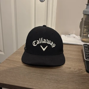 Svart Callaway keps Chrome Tour - Säljer en svart keps från Callaway med broderad logga framtill och texten 'Chrome Tour' på sidan. Baksidan har Odyssey-brodyr och sidan har även ELYTE-brodyr. Kepsen har klassisk form och justerbar rem baktill. Perfekt för golfbanan eller sportiga dagar.