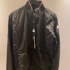 Svart vindjacka från Moncler - Säljer en svart vindjacka från Moncler med dragkedja framtill och klassisk krage med röd och vit rand. Jackan har Moncler-logga på ärmen och är tillverkad i lätt polyester. Perfekt för dig som vill ha en stilren och sportig look.