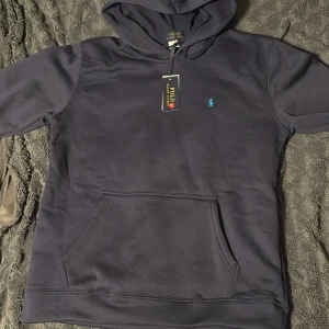 Svart hoodie från Polo Ralph Lauren - Svart hoodie från Polo Ralph Lauren med klassisk blå broderad logga på bröstet. Tröjan har huva med snörning och en stor magficka framtill. Tillverkad i mjuk bomull, perfekt för chill dagar. Snygg och enkel att matcha med allt.