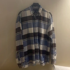 Rutig skjorta Tommy Hilfiger blå/brun - Snygg rutig skjorta från Tommy Hilfiger i blått, brunt och vitt. Skjortan har klassisk krage, långa ärmar och knappar framtill. Custom fit-modell som sitter riktigt bra och är perfekt för dig som gillar en stilren men avslappnad look. Går ner i pris om de önskas 