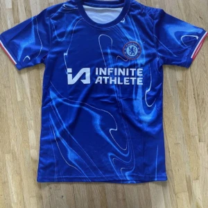 Chelsea blå fotbollströja Infinite Athlete - Snygg blå Chelsea fotbollströja med vit och röd detalj på ärmsluten. Tryck med Infinite Athlete på bröstet och Chelsea-logga. Tröjan har ett unikt abstrakt mönster i olika blå toner och är tillverkad i lätt polyester. Perfekt för dig som vill representera ditt lag.