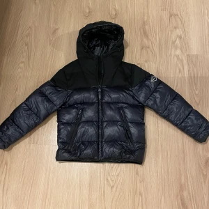 Svart pufferjacka från Replay - Säljer nu min vinterjacka då den knappt kommer till andvändning. Det är en glossy pufferjacka från Replay med huva och feta dragkedjor. Jackan har två sidofickor och två innerfickor med dragkedja samt en Replay-logga på armen. Jackan är perfekt för kalla dagar.  nypris ca 3600. Priset är ej hugget i sten, om det finns några frågor är det bara att skriva