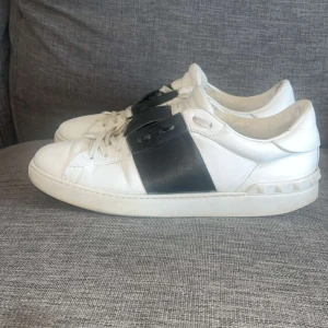 Valentino - Snygga Valentino Garavani Open sneakers i vitt skinn med bred svart läderdetalj över snörningen. Klassisk rund tå, platt sula och ikoniska nitar baktill. Perfekta för dig som gillar stilrena och lyxiga sneakers med en twist. Hör av dig vid frågor!