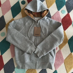 Grå hoodie från Burberry med dragkedja - Snygg grå hoodie från Burberry med klassiskt rutigt foder i huvan. Tröjan har dragkedja framtill, snörning i huvan och två fickor. Tillverkad i mjuk bomull och har långa ärmar. Perfekt för dig som vill ha en stilren och lyxig vardagslook. Lite liten i storlek så passar nog mera en medium. Helt ny bara provad säljes för 699kr.