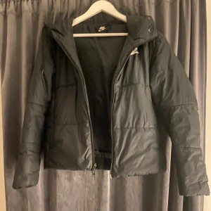 Svart pufferjacka från Nike - Svart pufferjacka från Nike med huva och dragkedja framtill.  Små Nike märken finns på insidan av luvan i en rad. Det fanns även en remsa Nedantill med nike märken men den är upplimmad med tyglim. Osäker på storlek men passar xs