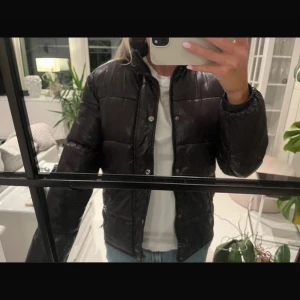 Svart pufferjacka från Guess - Svart pufferjacka från Guess med klassisk logga i rött och vitt på ryggen. Jackan har hög krage, tryckknappar och dragkedja framtill. Materialet är glansigt och känns riktigt mjukt. Perfekt för kalla dagar när du vill ha en stilren look. Kan vara något avskav av trycket bak men de ser man ej 