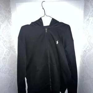 Polo Ralph Lauren Zip Hoodie - Svart hoodie från Ralph Lauren med dragkedja framtill och klassisk vit logga på bröstet. Tröjan har huva och känguruficka, perfekt för en chill och stilren look. Passar dig som gillar enkel och tidlös streetstyle.