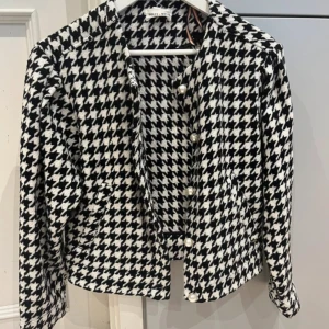 Rutig kavaj från Holly & Whyte XS - Snygg svartvit rutig kavaj från Holly & Whyte i storlek XS. Jackan har klassiskt hundtandsmönster, pärlknappar framtill och är långärmad. Materialet är mjukt och känns lyxigt, perfekt för att lyfta vilken outfit som helst.