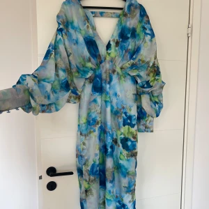 Blommig långklänning med v-ringning - Långklänning med v-ringning och stora, transparenta ärmar. Klänningen har ett blått och grönt blommigt mönster och är tillverkad i ett lätt, flygigt material. Perfekt för dig som vill sticka ut med en färgstark och elegant look.