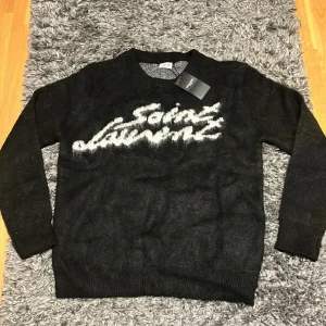 Svart Saint Laurent stickad tröja - Svart stickad tröja från Saint Laurent med vit logotyp framtill i en cool, lite rufsig stil. Tröjan har rund halsringning och långa ärmar. Tillverkad i 100% bomull för en skön känsla och snygg passform. (Storlek S men passar mer som en L)