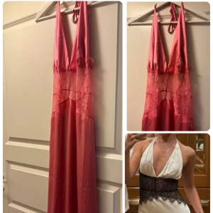 Rosa långklänning med spets och djup v-ringning - Snygg långklänning i rosa satin med djup v-ringning och tunna axelband som knyts i nacken. Klänningen har transparent spetsdetalj över midjan och en öppen rygg. Materialet är glansigt och faller mjukt, perfekt för dig som vill sticka ut.