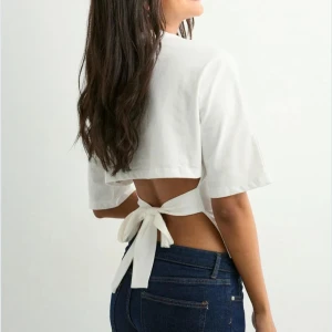 Vit topp med knyt i ryggen - Säljer en vit croppad topp med korta ärmar och snygg öppen rygg med knytband. Toppen har en enkel och stilren design med rund halsringning och är tillverkad i mjuk bomull. Perfekt för dig som vill ha en trendig och luftig look.