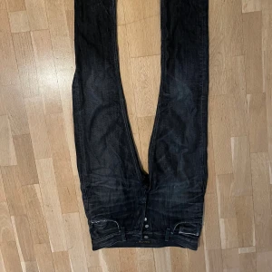 Svarta jeans från Nudie Jeans ovanlig färg  - Säljer ett par svarta jeans från Nudie Jeans med klassisk femficksdesign och snygga kontrastsömmar på bakfickorna. Jeansen är tillverkade i 100% ekologisk bomull och har en rak passform med knappgylf. Perfekta för dig som gillar stilrena och tidlösa jeans. Sample grim tin modell 