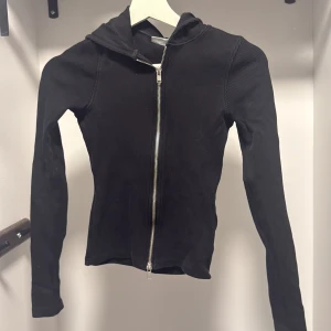 Svart ribbad hoodie från Weekday XS - Svart ribbad hoodie från Weekday i storlek XS. Tröjan har dragkedja framtill, huva och är långärmad. Snygg och enkel modell som sitter tight och passar perfekt till lager-på-lager-stil.