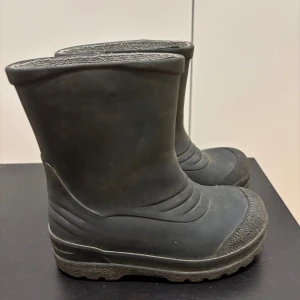 Svarta gummistövlar - Svarta gummistövlar med grov sula och rund tå. Klassisk modell i helsvart, perfekt för regniga dagar och leriga äventyr. Tillverkade i slitstarkt syntetmaterial med mönstrad yttersula för extra grepp.