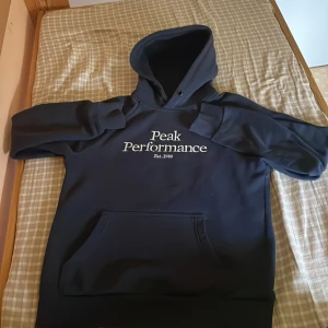 Mörkblå hoodie från Peak Performance - Snygg mörkblå hoodie från Peak Performance med vit logga på bröstet. Klassisk känguruficka framtill, huva och ribbade muddar. Perfekt för chill dagar eller när du vill ha en clean och sportig look.