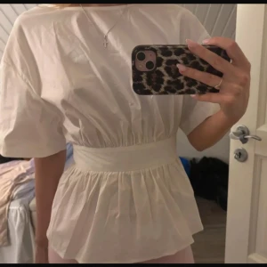 Vit blus med öppen rygg och rosett - Säljer en vit blus med korta puffärmar och bred midjedetalj. Blusen har en snygg öppen rygg med stor rosett som knyts baktill. Materialet är lätt och känns fräscht, perfekt för dig som gillar en feminin och trendig look.