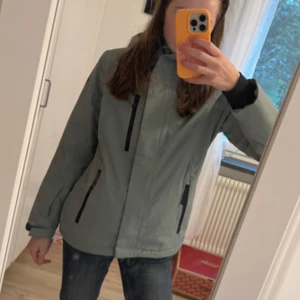  skidjacka - Snygg ljusgrön skidjacka med svart foder och huva. Jackan har tre svarta dragkedjefickor framtill, justerbara ärmslut och en stilren look. Perfekt för dig som gillar enkel och clean stil. Passformen är normal och jackan är tillverkad i syntetmaterial.