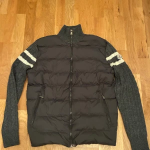 Grå Moncler pufferjacka med stickade ärmar - Snygg pufferjacka från Moncler i mörkgrått med stickade ärmar och krage. Ärmarna har vita ränder och Moncler-logga på vänster arm. Jackan har dragkedja framtill och två fickor med dragkedja. Perfekt för kyliga dagar när du vill ha både stil och komfort.