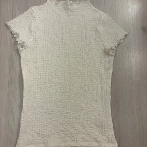 Vit smockad topp från Kappahl - Supersöt vit smockad topp från Kappahl med korta ärmar och volangkant vid hals och ärmslut. Toppen har en tight passform och är gjord i ett stretchigt material som sitter snyggt på kroppen. Perfekt att styla med jeans eller kjol.
