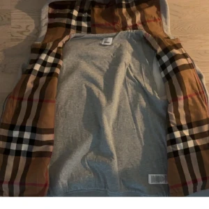 Grå hoodiejacka med rutig huva Burberry - Snygg grå hoodiejacka från Burberry med dragkedja och två fickor framtill. Insidan av huvan har det klassiska Burberry-rutiga mönstret i brunt, svart, vitt och rött. Perfekt för dig som vill ha en stilren jacka med exklusiv touch.