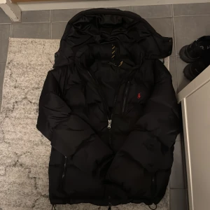 Svart pufferjacka Polo Ralph Lauren M - Svart pufferjacka från Polo Ralph Lauren med huva, dubbla dragkedjor och RL-detaljer på både dragkedja och justerbara ärmslut. Jackan har klassisk röd Polo-logga på bröstet och är tillverkad i polyester samt har en dunfyllning. Luvan är bortagbar och vill sälja då M inte passar utan behöver L. Pris kan diskuteras vid snabb affär. Använd ca 8 gånger innan den kändes för kort.