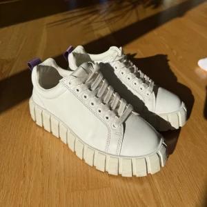 Chunky vita sneakers från Eytys - Säljer ett par chunky vita sneakers från Eytys med kraftig plattformsula och lila hälflik. Skorna är i skinn och har snörning framtill samt rund tå. Perfekta för dig som gillar statement-sneakers med cool design och extra höjd. Säljer då de är för små, använda extremt fåtal gånger. Nypris 2200kr