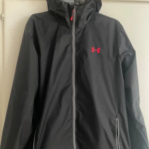 Svart vindjacka från Under Armour M - Svart vindjacka från Under Armour med röd logga på bröstet och huva. Jackan har hel dragkedja framtill, två fickor med dragkedja och meshfoder på insidan. Perfekt för blåsiga dagar och sportiga outfits.