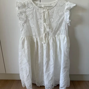 Vit broderad kortklänning med volang - Superfin vit kortklänning med broderade blommor, volangdetaljer på axlarna och knytning framtill. Klänningen är ärmlös och har en luftig, romantisk känsla – perfekt för varma dagar. Tillverkad i bomull för en skön och sval känsla.