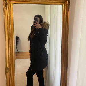 Rock and blue jacka  - Svart lång Rock and blue jacka storlek 32 med stor huva och fluffig äkta päls i beige runt kragen. Jackan har dragkedja, knappar, fickor med lock och en extra ficka med dragkedja på ärmen. Perfekt för kalla vinterdagar och riktigt snygg siluett - ordinarie pris 5000