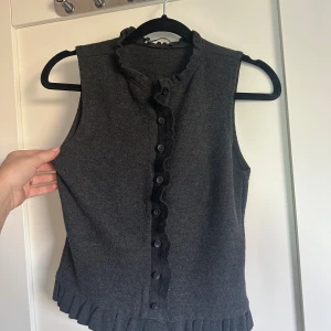 Mörkgrå stickad väst med volang - Mysig mörkgrå stickad väst med knappar framtill och volangdetaljer längs knappraden och nederkanten. Säljer även o beige.