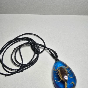 Halsband med blå skorpionhänge - Unikt halsband med svart flätad snodd och ett droppformat hänge i blå resin med en riktig skorpion inuti. Perfekt för dig som gillar annorlunda och coola accessoarer. Hänget har en intensiv blå färg som verkligen sticker ut.