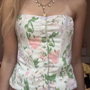Blommig korsett i pastellfärger - Superfin korsett med blommigt mönster i ljusa pastellfärger som rosa, grön, lila och vit. Toppen är axelbandslös och har en figurnära passform med markerade sömmar framtill. Perfekt för dig som vill ha en romantisk och trendig look.