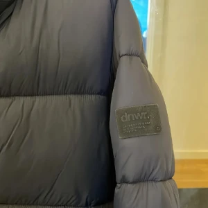 Svart pufferjacka från dnwr - Säljer en svart pufferjacka från dnwr med hög krage och breda, quiltade sektioner. Jackan har dragkedja framtill och två sidofickor med dragkedja. Märkespatch på ärmen ger en cool streetkänsla. Perfekt för kalla dagar och riktigt snygg till jeans.