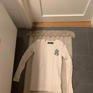 Vit kabelstickad tröja från Lauren Ralph Lauren - Kabelstickad vit tröja från Lauren Ralph Lauren med blå broderad logga på bröstet och rund halsringning.(Och den är äkta.) Tröjan har långa ärmar, ribbade muddar och dekorativa knappar på axeln. Perfekt för en klassisk och stilren look.