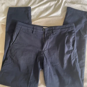 Mörkblå straight chinos från Selected Homme - Snygga mörkblå chinos från Selected Homme i rak modell. Byxorna har klassiska sidfickor, bälteshällor och en bakficka med knapp. Materialet är mjukt och lättskött, perfekt för en clean och stilren look. 