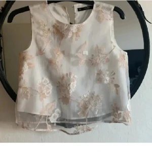 Zara Blommig ärmlös blus i ljusa toner - Superfin ärmlös blus från Zara i transparent vitt tyg med broderade blommor i beige och ljusrosa. Blusen har rund halsringning och lätt boxig passform, perfekt för en fräsch och somrig look. Snygg att styla med både jeans och kjol.