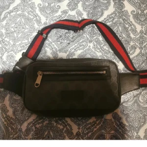 Gucci Väska - Cool svart axelväska med diskret mönster och en dragkedja framtill. Väskan har en bred, justerbar rem i rött och mörkblått som ger en sportig vibe. Perfekt för att bära det viktigaste och hålla händerna fria.