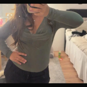 Olivgrön långärmad body med v-ringning - Snygg olivgrön långärmad body med djup v-ringning och draperad detalj framtill. Bodyn har en tight passform och är tillverkad i ett mjukt, stretchigt material som sitter skönt på kroppen. Perfekt att styla med jeans eller kjol för en trendig look.