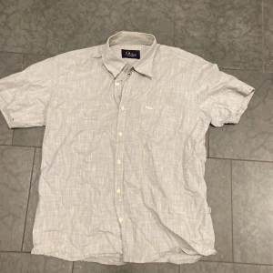 Grå kortärmad Peak Performance skjorta - Kortärmad skjorta från Peak Performance i ljusgrått med diskret rutigt mönster. Klassisk krage, knäppning framtill och liten bröstficka med broderad logga. Perfekt för en chill och stilren look.